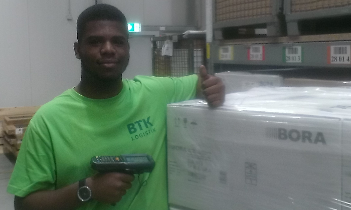 Brandon Ozomoru, der erste Auszubildende im Logistikzentrum der BTK in Raubling, steht mit einem Scanner in der Hand an einer Palette mit Waren von Bora. Es ist sein erster Ausbildungstag.